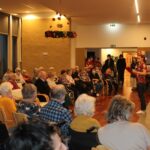 Stanovalci doma starejših sedijo v dvorani in spremljajo praznično prireditev z nastopajočo, ki bere besedilo.