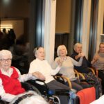 Skupina starejših stanovalcev sedi v krogu, se drži za roke in sodeluje v skupni dejavnosti.