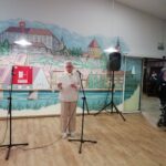 Starejša gospa stoji za mikrofonom in bere besedilo na praznični prireditvi v dvorani.