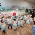 Otroci v belih in svetlo modrih oblekah nastopajo s plesom v dvorani, vzgojiteljica kleči pred njimi in jih usmerja.