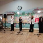 Štiri pevke stojijo pred mikrofoni in nastopajo na odru ob božično okrašeni dvorani.