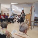 Ženska pripovedovalka stoji pred skupino starejših oseb, ki sedijo v krogu v invalidskih vozičkih in stolih ter poslušajo predstavitev. Ob njej stoji leseno stojalo za kamišibaj.