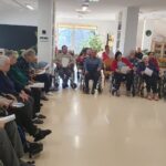 Večja skupina starejših oseb sedi v polkrogu, nekateri v invalidskih vozičkih, in posluša pripovedovanje. V ospredju je kamišibaj stojalo s sliko zimskega drevesa.