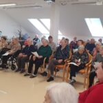 Skupina starejših oseb sedi v vrsti stolov in invalidskih vozičkov. Nekateri držijo list papirja, prostor je svetel z velikimi strešnimi okni.