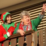 Dve osebi stojita na balkonu; ena oseba je oblečena v praznični kostum, druga pa maha proti dogajanju spodaj.