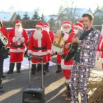 Skupina oseb na prostem stoji ob parkirišču; več oseb je oblečenih v rdeče praznične kostume, ena oseba igra kitaro.