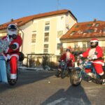 Več oseb v rdečih prazničnih kostumih se pripelje z mopedi pred stavbo; v ozadju so vidni balkon, parkirana vozila in vhod v stavbo.
