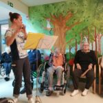 Oseba stoji ob notnem stojalu in igra flavto, več oseb sedi ob steni in posluša.