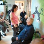 Oseba v delovni uniformi se pogovarja z osebo na invalidskem vozičku, druge osebe sedijo ob hodniku.