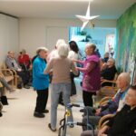 Več oseb stoji v krogu in se drži za roke med plesom, druge osebe sedijo ob stenah in opazujejo.