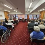 Več oseb sedi na stolih v dvorani in spremlja glasbeni nastop otrok z harmonikami spredaj.