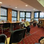 Otrok sedi na stolu in igra harmoniko, v ozadju so drugi otroci z glasbili in odrasle osebe.