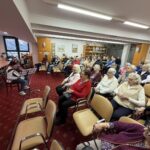 Otrok sedi pred notnim stojalom in igra harmoniko, občinstvo sedi v vrstah in posluša nastop.