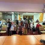 Skupina otrok stoji spredaj in drži harmonike, za njimi sedi občinstvo v dvorani.