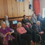 Skupina oseb sedi na stolih in foteljih v skupnem prostoru ter gleda v isto smer, ob nekaterih so pripomočki za hojo.