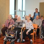 Skupina oseb sedi v večnamenskem prostoru, nekatere uporabljajo pripomočke za hojo ali invalidske vozičke, ena oseba stoji za njimi.