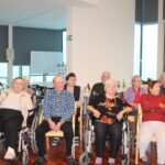 Več oseb sedi v večnamenskem prostoru, razporejene so v vrstah, vse gledajo v isto smer.