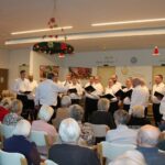 Več oseb sedi v večnamenskem prostoru in spremlja nastop moškega pevskega zbora, ki stoji v polkrogu pred njimi.