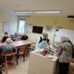 Več oseb sedi okoli mize v prostoru, dve osebi pri pultu pripravljata hrano na kuhalni plošči.