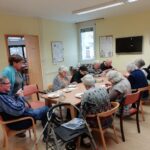 Več oseb sedi okoli mize in je sladico s krožnikov, na mizi so krožniki in pribor.