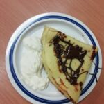 Krožnik z zloženo palačinko, prelito s čokolado, in kepo bele kreme na lesenem ozadju.