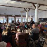 Več oseb sedi za mizami v večjem skupnem prostoru in se pogovarja, ob mizah so tudi invalidski vozički, v ozadju stojijo druge osebe.