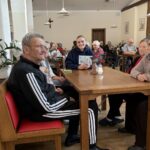 Skupina oseb sedi okoli mize, ena oseba sedi na invalidskem vozičku, druga oseba drži list z napisom, prostor je svetel in urejen.