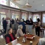 Več oseb stoji in sedi v skupnem prostoru, ena oseba igra na harmoniko, druge osebe stojijo skupaj in spremljajo dogajanje.