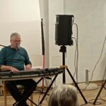 Oseba sedi za električnimi klaviaturami in poje v mikrofon, ob njem sta zvočnika na stojalih, v ospredju so vidne glave poslušalcev.