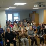 Skupina oseb sedi v več vrstah na stolih in invalidskih vozičkih ter gleda proti odru v skupnem prostoru.