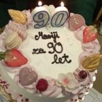 Okrašena torta z dvema prižganima svečkama v obliki številke 90, okrašena s sadjem, cvetovi in čokoladnim napisom.