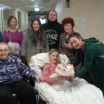 Skupina oseb stoji in sedi okoli osebe v postelji, ena oseba drži torto s prižganimi svečkami.