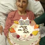 Oseba sedi v postelji in gleda torto s prižganimi svečkami v obliki številke 90, torto držijo roke druge osebe.