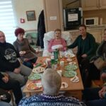 Več oseb sedi okoli mize s krožniki sladice in pijačo, ena oseba sedi v postelji ob mizi v prostoru, ki spominja na skupni prostor.