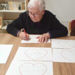 Oseba sedi za mizo in piše z rdečim svinčnikom na list papirja z narisanim srcem.