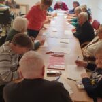 Več oseb sedi okoli dolge mize in piše na papir z narisanimi srci, ena oseba stoji in pomaga.