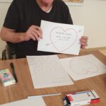 Oseba sedi za mizo in drži list papirja z narisanim srcem in rokopisnim sporočilom, na mizi so pisala in drugi listi.