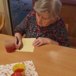 Stanovalka doma za starejše sedi za mizo in piše novoletno voščilo na list papirja.