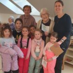 Skupina otrok in starejših oseb stoji in sedi skupaj v prostoru, ena oseba drži voščilnico, več otrok stoji v prvi vrsti.