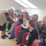 Več otrok sedi na klopi, starejša oseba jim ponuja košaro z vsebino, ostale osebe sedijo v ozadju.