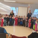 Skupina otrok stoji v vrsti v večnamenskem prostoru, odrasli osebi stojita ob strani, starejše osebe sedijo okoli in opazujejo.