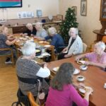 Širši pogled na skupni prostor, kjer več oseb sedi za dvema mizama in je palačinke, na mizah so krožniki in skodelice, v prostoru je vidna kuhinjska oprema.