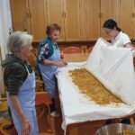 Stanovalke in zaposlena skupaj pripravljajo zavitek; na raztegnjeno testo enakomerno razporejajo nadev.