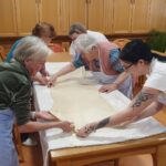 Skupina stanovalcev in zaposlena raztegujejo testo za zavitek na veliki mizi v skupnem prostoru.