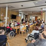 Več oseb sedi za mizami v velikem skupnem prostoru, nekatere so na invalidskih vozičkih, v ospredju je viden nastop glasbenikov.
