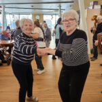 Dve osebi stojita v skupnem prostoru in se držita za roke, kot pri plesu, okoli njiju so druge osebe in glasbeniki.