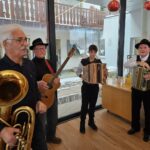 Štiri osebe stojijo v notranjem prostoru in igrajo na glasbila, med njimi so trobilo, kitara in harmonike.