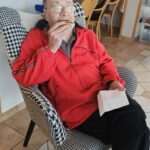 Oseba sedi v naslonjaču in drži kos kruha, v ozadju pa je viden skupni prostor z drugimi osebami, ki sedijo za mizo.