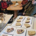 Več oseb sedi za mizo v skupnem prostoru, pred njimi so kozarci in na mizi so postavljeni krožniki, v ospredju pa je voziček z narezanimi kosi kruha.