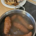 Na kuhalni plošči stoji lonec z vodo, v katerem se kuhajo klobase, ob njem pa je na pultu krožnik z narezanimi rezinami peciva.